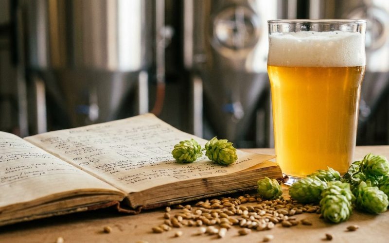Cuaderno de recetas con malta y lúpulo frente a tanques de cervecería